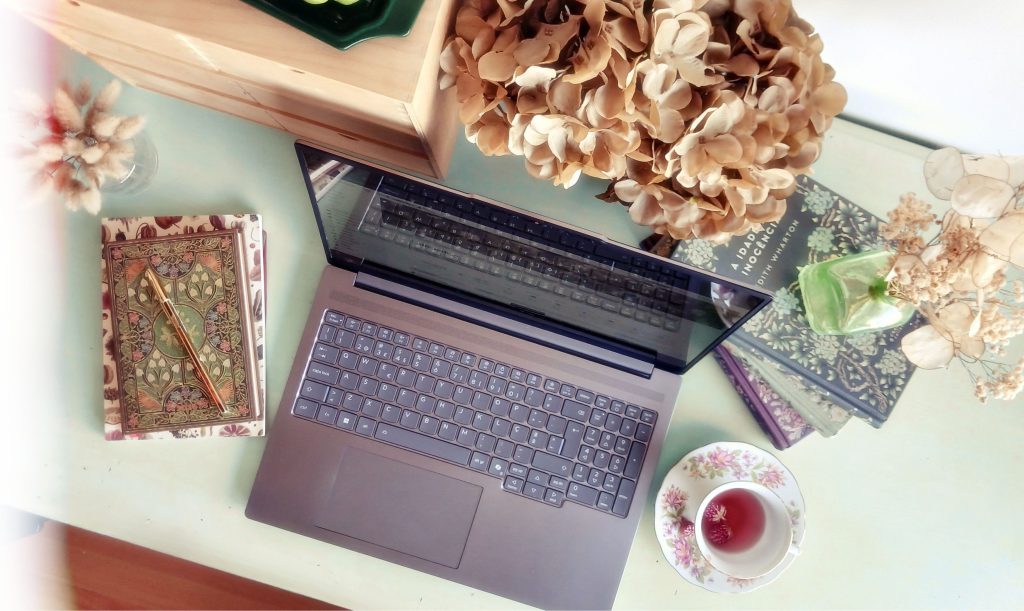 Plano de cima de uma secretária organizada com portátil, flores, bloco de notas e canetas, espaço de trabalho de uma assistente virtual, estilo cottagecore ou light academia