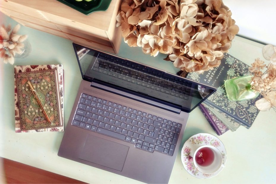 Plano de cima de uma secretária organizada com portátil, flores, bloco de notas e canetas, espaço de trabalho de uma assistente virtual, estilo cottagecore ou light academia