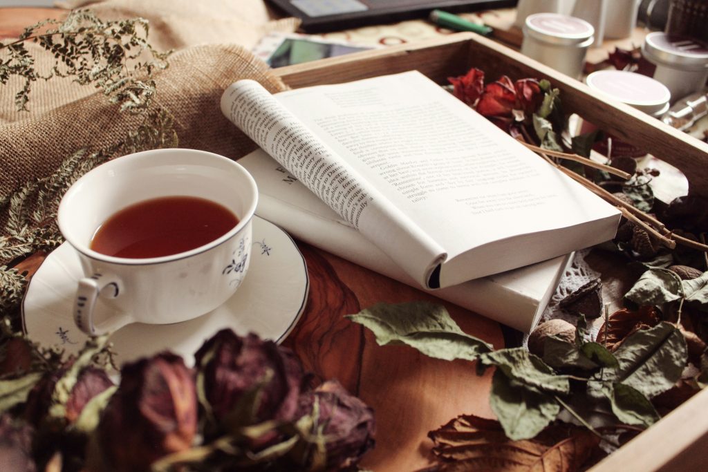 Livro aberto ao lado de uma chávena de chá vintage num tabuleiro, rodeado de flores secas — pausa criativa e inspiração para empreendedoras criativas